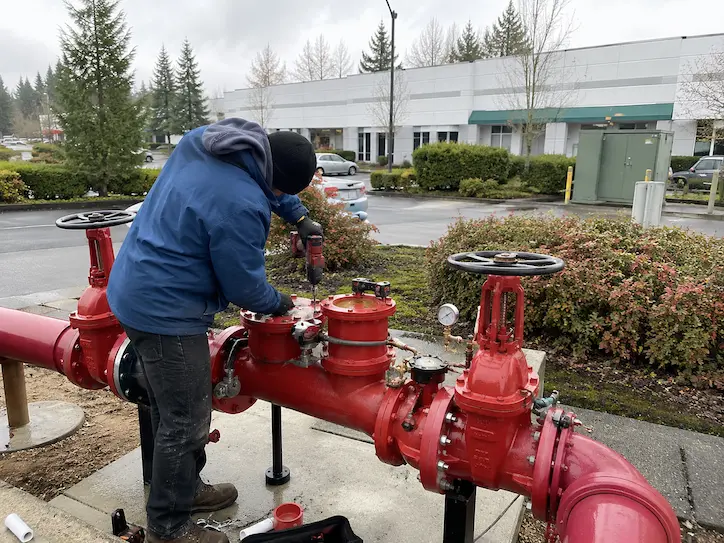 Commercial backflow testing technician servicing a large red backflow prevention assembly outside a Northwest commercial facility for Elite Mechanical.