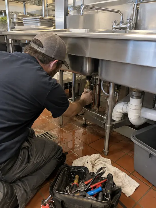 Plumber inspecting supply and drain lines beneath a commercial kitchen sink